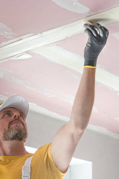 The worker make a plasterboard ceiling. He does taping plaster drywall ceiling joints. He is inserting the glass tape into the putty.